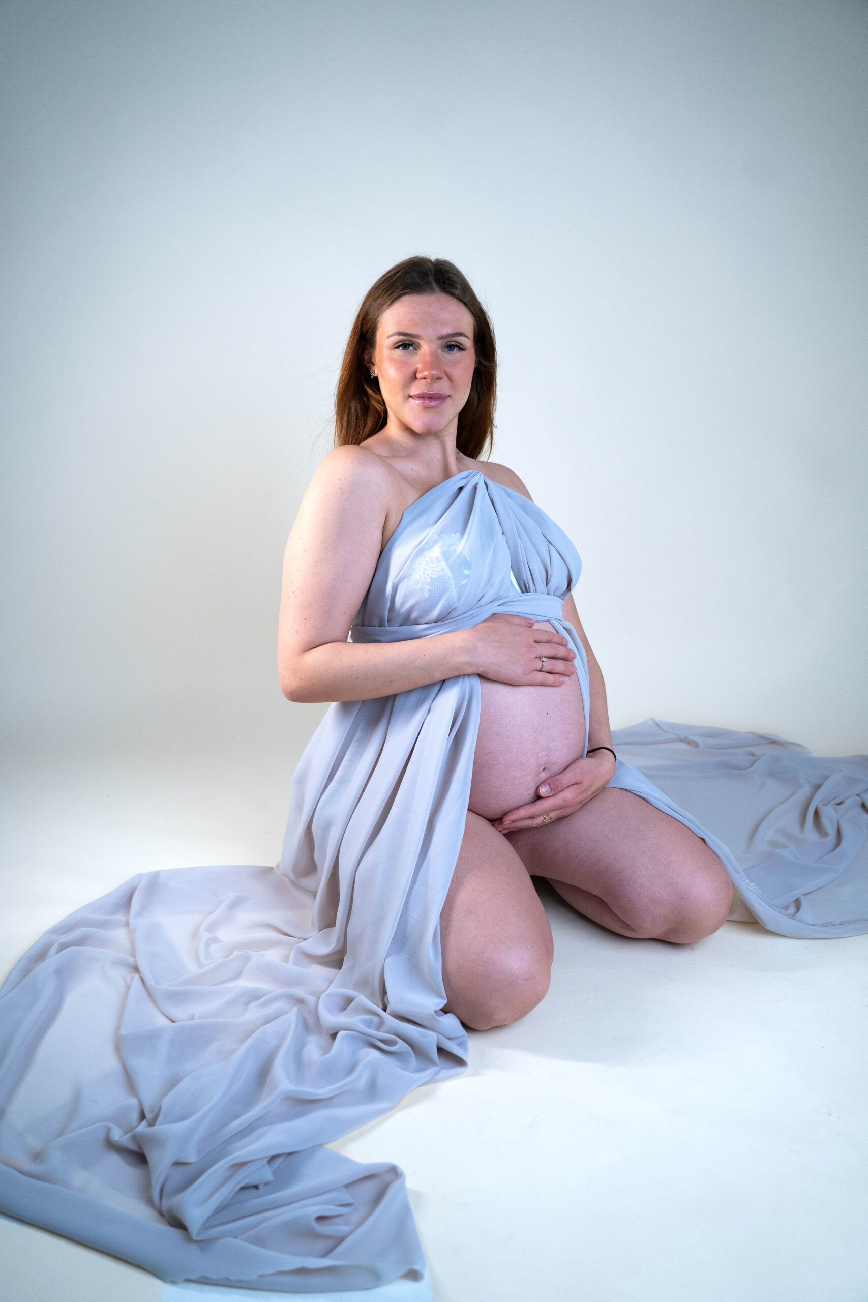 Schwangere Frau mit Babybauch bei einem Schwangerschaftsshooting sitzend in elegantem langen Kleid